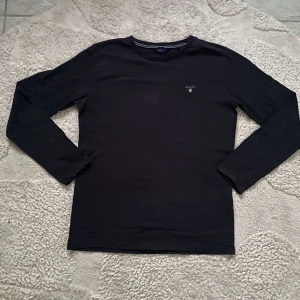 Mörkblå långärmad tröja från GANT - Säljer en stilren mörkblå långärmad tröja från GANT med broderad logotyp på bröstet. Perfekt för en avslappnad och snygg look. Tröjan är i en klassisk passform och tillverkad i mjukt material.