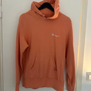 Orange hoodie från Champion - Säljer en snygg orange hoodie från Champion. Den har en klassisk känguruficka och ribbade muddar. Perfekt för en avslappnad stil. Champion-loggan syns både på bröstet och ärmen.