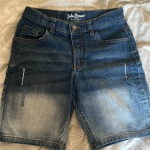 Jeansshorts från John Baner för barn! - Snygga jeansshorts från John Baner i en klassisk blå denimfärg. De har en normal passform med fem fickor och en knappgylf. Perfekta för en avslappnad stil. Märkeslogga på baksidan ger en extra touch.