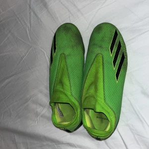 Gröna fotbollsskor från Adidas - Snygga gröna fotbollsskor från Adidas med svarta detaljer. Skorna har en strömlinjeformad design utan snörning och är perfekta för snabba rörelser på planen. Yttersulan har flera dobbar för bra grepp.