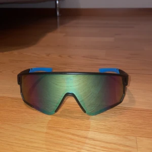 Sportiga solglasögon från MaxAir - Snygga sportiga solglasögon från MaxAir med en spegelreflekterande lins i regnbågsfärger. Bågarna är svarta med blå detaljer på skalmarna. Perfekta för en aktiv livsstil och ger ett coolt intryck. Riktigt snygga