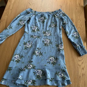 Blommig offshoulder klänning från H&M Divided - Säljer en söt ljusblå offshoulder klänning från H&M Divided med blommigt mönster. Klänningen har långa ärmar med elastiska muddar och volangdetaljer. Den är i strl 40 men passar även för 38, och blir aningens oversized för 36 men mer med ett ”svall” isåfall. Dragkedja i midjan!