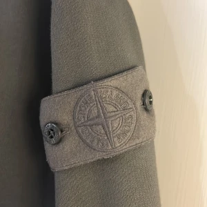 Grå jacka från Stone Island - Säljer en oanvänd grå tröja från Stone Island, storlek L, med dragkedja, de gråa märket på armen är unikt!