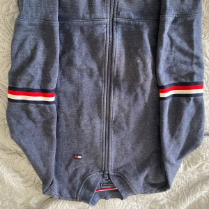 Grå hoodie från Tommy Hilfiger - Säljer en stilren grå hoodie från Tommy Hilfiger med dragkedja och ribbade muddar. Tröjan har märkets ikoniska ränder i rött, vitt och blått på ärmarna och en liten logotyp på framsidan. Perfekt för en avslappnad look.