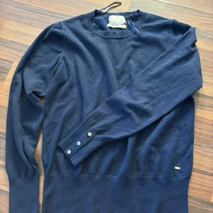 Mörkblå tröja från Holly & whyte - Säljer en stilren mörkblå tröja från Holly & whyte med långa ärmar och knappar vid ärmsluten. Tröjan har en rund halsringning och en liten metallbricka med H&W-logga vid nederkanten. Perfekt för en klassisk look.