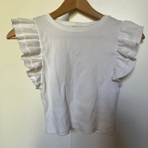 Vit ribbad topp från Zara - Säljer en söt vit ribbad topp från Zara med volangdetaljer på ärmarna. Perfekt för en stilren och trendig look. Toppen är ärmlös och har en bekväm passform. Storlek s men passar även xs