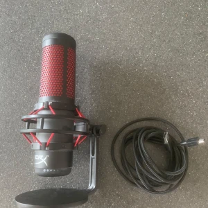 HyperX QuadCast mikrofon - Säljer en HyperX mikrofon i svart och rött med USB-anslutning. Perfekt för gaming eller streaming. Inkluderar stativ och USB-kabel. Mikrofonen har en modern design med ett rött nätmönster och svart bas.