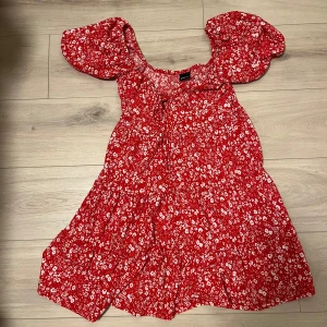 Röd blommig klänning från Gina Tricot  - Charmig röd klänning med vita blommor och puffiga kortärmar från Gina Tricot. Perfekt för sommarens varma dagar. Den har en söt och luftig design som ger en romantisk känsla.