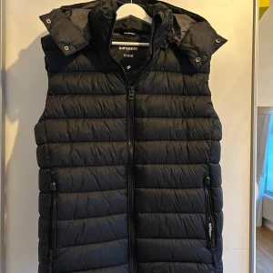 Superdry väst - Svart dunväst från Superdry med dragkedja och avtagbar huva. Storlek L, skicket är som nytt.
