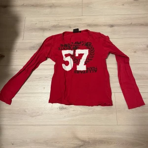 Röd långärmad tröja med tryck - Säljer en röd långärmad tröja med ett stort vitt nummer 57 tryckt på framsidan. Tröjan har en avslappnad passform och är perfekt för en casual look. Passar bra till jeans eller shorts.
