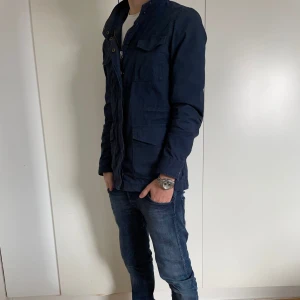 Mörkblå fieldjacka - Säljer en stilren mörkblå field jacka med dragkedja och knappar. Jackan har flera praktiska fickor och en hög krage. Perfekt för en casual look. Skick 8.5/10 Modellen är 173 Passar xs-s