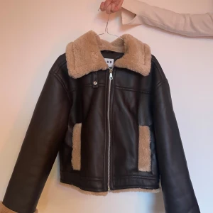 Brun ”skinnjacka” med foder från NA-KD - Snygg brun jacka i bondat material från NA-KD med mjukt beige foder. Jackan har en dragkedja framtill och en fluffig krage. Tyvärr inte kommit till användning. Använd 2-3 gånger. 
