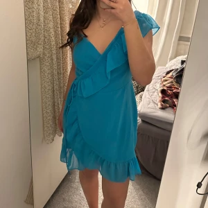 Blå omlottklänning - Säljer en snygg blå omlottklänning med volangdetaljer. Klänningen har en v-ringad design och kort längd, perfekt för sommarens fester. Den är ärmlös och har ett knytband i midjan för en justerbar passform. Perfekt för att känna sig både bekväm och stilren.