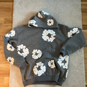 Grå hoodie med vita blommor från Denim Tears - Säljer en grå hoodie från Denim Tears med vita blommönster över hela tröjan. Den har en klassisk passform med huva och långa ärmar. Perfekt för en avslappnad stil. Det går att få ner priset lite!!vid frågor skriv!🤝