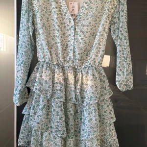 Blommig klänning med volanger - Säljer en söt blommig klänning med volanger i ljusgrönt och vitt. Klänningen har långa ärmar och en v-ringad design med knappar framtill. Perfekt för vår och sommar! Från Gina 💘Passar mig som är S