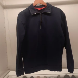 Mörkblå tröja med dragkedja från RYVLS - Säljer en stilren mörkblå tröja från RYVLS med dragkedja i halsen. Perfekt för en avslappnad look. Tröjan har långa ärmar och en hög krage som kan justeras med dragkedjan. Passar bra till både jeans och chinos.