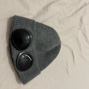 Grå C.P Company Mössa   - Säljer en stilren och trendig C.P. Company Goggle Beanie i grå färg. Mössan är i bra skick och perfekt för både streetwear och casual outfits. 	•	Material: Högkvalitativ stickad bomullsblandning 	•	Färg: Grå 	•	Detaljer: Ikoniska goggles på uppviket 	•	Passform: One size
