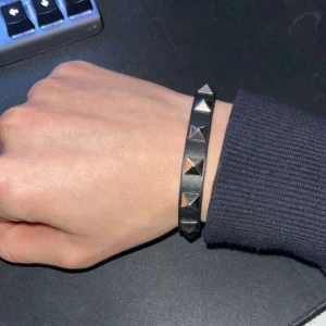 Svart armband med nitar - Snyggt svart armband med silverfärgade pyramidnitar. Armbandet har ett spänne i guldton för justerbar passform. Perfekt för att ge en edgy touch till din stil.