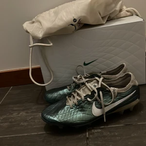 Nike fotbollsskor i grönt och vitt - Snygga fotbollsskor Nike tiempo legend 10 elite i en metallicgrön färg med vita detaljer. Skorna har snörning och en quiltad yta för extra stil. Perfekta för fotbollsplanen med sina robusta dobbar. Nypris 2000