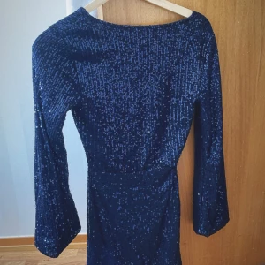 Mörkblå glittrig klänning - Snygg mörkblå klänning med glittriga paljetter som ger en festlig look. Klänningen har långa ärmar, lite öppen rygg och en kort längd, perfekt till glitterfest eller nyår