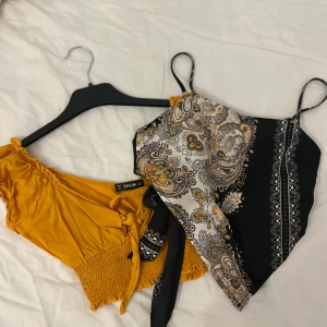 Mönstrad och gul topp - paketpris  - Ena är en mönstrad linne med paisley-mönster i svart, vitt och gult, med smala axelband och knytning fram. Den andra är en senapsgul offshoulder-topp med korta ärmar och smockdetaljer i midjan. 1st för 50kr BÅDA FÖR 80kr