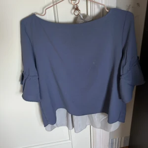 Blå blus med volangärmar - Snygg blå blus med trekvartsärm och volangdetaljer. Blusen har en unik omlott-design i ryggen som ger en stilfull look. Perfekt för att piffa upp garderoben!