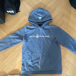 Blå hoodie från Sail Racing - Säljer en blå hoodie från Sail Racing med vit logga på bröstet. Tröjan har en stor ficka framtill och en bekväm huva. Perfekt för en avslappnad stil.