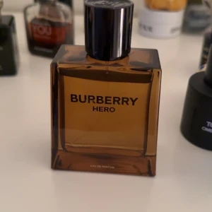 Burberry Hero Eau de Parfum - Säljer en elegant Burberry Hero Eau de Parfum i en stilren brun glasflaska med svart lock. Doften är maskulin och modern, perfekt för den som vill ha en sofistikerad touch. Flaskan har en fyrkantig form och rymmer en doft som utstrålar självsäkerhet.