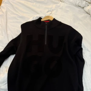 Svart tröja med dragkedja från Hugo - Snygg svart tröja från Hugo med stor logotyp framtill. Tröjan har en dragkedja vid halsen och långa ärmar. Perfekt för en stilren look.