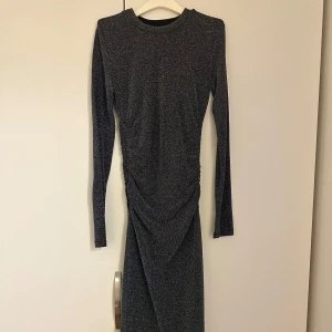 Glittrig långärmad klänning - Snygg glittrig klänning med långa ärmar och en elegant drapering framtill. Perfekt för en kväll ute eller en speciell tillställning. Klänningen har en smickrande passform och är i en mörk färgton.