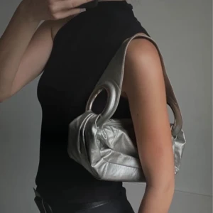 Silver axelväska med ringdetaljer - Snygg silverfärgad axelväska i syntetmaterial med coola ringdetaljer. Perfekt för en utekväll eller fest! Väskan har en unik design som verkligen sticker ut. Den är både stilren och praktisk med en dragkedja för säker förvaring.