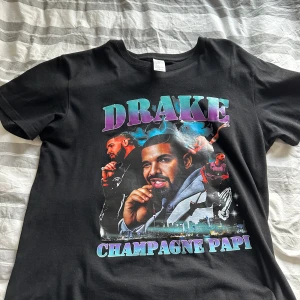 Svart t-shirt med Drake-tryck - Cool svart t-shirt med färgglatt tryck av artisten Drake och texten 'Champagne Papi'. Perfekt för fans av hans musik! T-shirten är kortärmad och gjord i mjukt material för en bekväm passform. Passar perfekt till vardags eller konsertbesök.