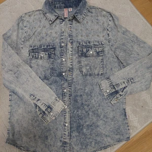 Acid wash jeansskjorta med stjärndetaljer - Säljer en cool acid wash/gråblå jeansskjorta från Wow Cubus i storlek S. Skjortan har långa ärmar och är dekorerad med små stjärnor på framsidan och har två bröstfickor med knappar. Skjortan är i bra skick men den har används några gånger, det finns inga deffekter eller annat.