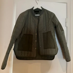 Grön quiltad jacka - Säljer en snygg grön quiltad jacka med knappar framtill. Den har långa ärmar och stora fickor som ger en cool look. Perfekt för höst och vår när du vill hålla dig varm men ändå se stilren ut. Passar till både vardag och lite finare tillfällen.