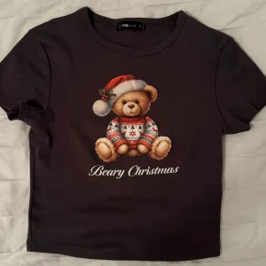 Svart jultröja med nallemotiv - Säljer en svart kortärmad tröja med ett gulligt nallemotiv som har på sig en jultröja och tomteluva. Texten 'Beary Christmas' står under nallen. Perfekt för julfirandet! 🎄