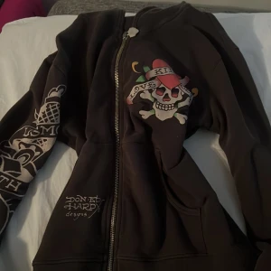 Grå hoodie från Ed Hardy - Säljer en cool brun hoodie från Ed Hardy med ikoniska dödskalle- och hjärtmotiv. Den har dragkedja och tryck på både fram- och baksida. Perfekt för en avslappnad stil med en edgy touch. Passar bra till höst och vår! De var ai som skrev allat INTE JAG plz köp så jag kan skaffa julpresenter till mina nära och kära den har inga defekter den är pawsome 