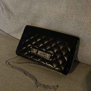 Svart quiltad axelväska från Love Moschino - Enbart använd enstaka gång. Snygg svart quiltad axelväska från Love Moschino med en elegant kedjerem. Perfekt för både vardag och fest. Väskan har en stilren design med logotypen i silver på framsidan. En chic accessoar för alla tillfällen!