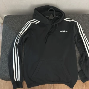 Svart hoodie från Adidas - Säljer en svart Adidas hoodie i storlek S. Den har de klassiska tre ränderna i vitt längs ärmarna och en stor ficka framtill. Perfekt för både träning och vardag. Huvan har dragsko för justering. Skön och stilren design!