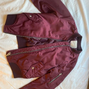 Vinröd bomberjacka från Pull&Bear - Säljer en snygg vinröd bomberjacka från Pull&Bear i storlek S. Jackan har en dragkedja framtill och ribbade muddar vid ärmslut och nederkant. Perfekt för höst och vår! 🧥