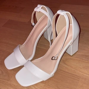 Vita klackskor med remmar - Snygga vita klackskor i syntetmaterial med en stilren design. De har en fyrkantig tå och breda klackar för extra stabilitet. Remmarna runt ankeln ger en elegant touch och säker passform. Perfekta för fest eller bal!