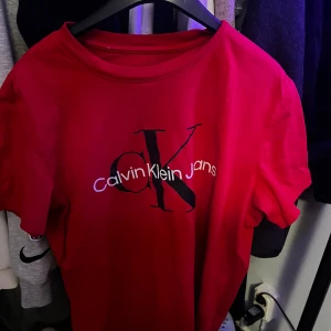 Röd t-shirt från Calvin Klein - Säljer en snygg röd t-shirt från Calvin Klein Jeans. Den har en klassisk passform med korta ärmar och ett stort CK-logotryck på framsidan. Perfekt för en casual look eller att styla upp med en kavaj. Passar både till vardags och fest!