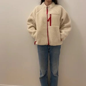 Beige teddyjacka från Moussy - Mysig beige teddyjacka från Moussy med röd dragkedja och fickdetaljer. Perfekt för kyliga dagar med sin varma och fluffiga design. Jackan har en normal passform och långa ärmar. En stilren och bekväm jacka för både höst och vinter.