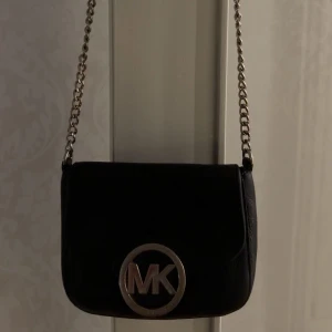 Svart axelväska från Michael Kors - Snygg svart axelväska från Michael Kors med en elegant kedjerem. Väskan har en stor MK-logga i silver på framsidan som ger en lyxig känsla. Perfekt för både vardag och fest! 🖤