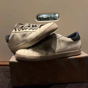 Golden Goose Superstar sneakers - Säljer ett par Golden Goose Superstar sneakers i vitt med grå stjärndetalj och mörkblå häl. Skorna har en cool, sliten look och är perfekta för en avslappnad stil. De har snörning och är i bra skick. Perfekta för både vardag och fest!