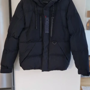 Svart dunjacka från Hoodrich - Säljer en svart dunjacka från Hoodrich i mycket bra skick. Jackan har en cool streetwear-stil med tryck på ryggen och insidan. Den är långärmad och har en dragkedja framtill. Perfekt för höst och vinter!