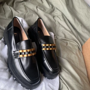 Svarta loafers med gulddetaljer från Zara - Snygga svarta loafers i skinn från Zara med coola gulddetaljer på ovansidan. De har en robust sula som ger bra grepp och en stilren look. Perfekta för både vardag och fest! Skorna är i mycket bra skick och redo att användas direkt.