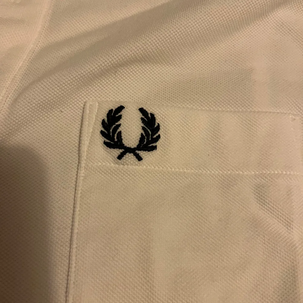 Säljer en klassisk vit pikétröja från Fred Perry i storlek M. Tröjan är i 100% bomull och har en broderad logga på bröstet. Perfekt för en stilren och avslappnad look. Den har korta ärmar och knappar vid kragen. Passar både till vardags och fest!. T-shirts.