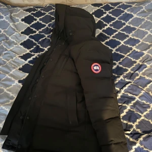 Svart dunjacka med pälsdetalj - Säljer en svart dunjacka från Canada Goose i mycket bra skick. Jackan har en avtagbar pälsdetalj på huvan och är perfekt för kalla vinterdagar. Den har en dragkedja och knappar framtill för extra skydd mot kyla. Supervarm och stilren!