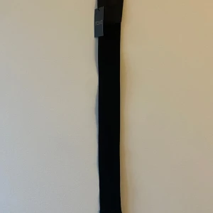 Svart bälte från Qnuz - Snyggt svart bälte från Qnuz med en unik rund spänne-design. Perfekt för att ge din outfit en stilren touch. Bältet är i nytt skick.