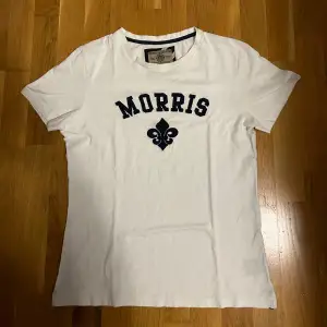 Säljer en snygg vit t-shirt från Morris i storlek S. Den är gjord av 100% bomull och har en cool text och symbol på bröstet. Perfekt för en avslappnad stil. T-shirten är i bra skick och redo för nya äventyr! Behöver bara strykas.